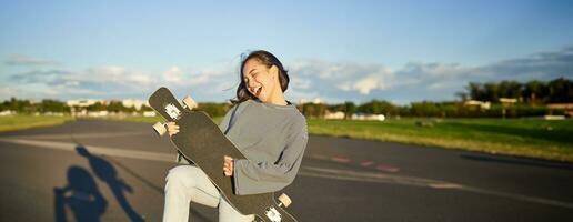 gracioso asiático niña disfrutando Patinaje, participación patineta me gusta guitarra y sombra jugando, teniendo divertido al aire libre foto