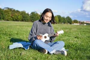 sonriente asiático niña mirando a guitarra aplicación, canto y jugando ukelele mientras echar un vistazo a acordes en teléfono inteligente, sentado al aire libre en parque en cobija foto