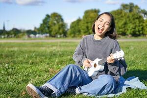 retrato de hermosa asiático mujer cantando, jugando ukelele guitarra en parque, sentado solo en césped en soleado día foto