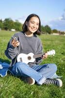 Vertical shot of smiling korean girl sitting in park with ukulele and smartphone, using mobile app to tune instrument, looking for chords while playing photo