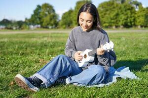 despreocupado asiático niña canto y jugando ukelele en parque, sentado en césped, músico relajante en su gratis hora al aire libre foto