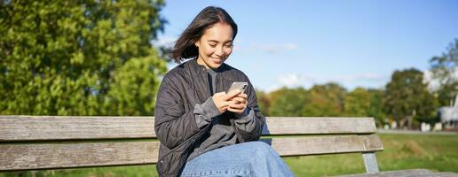 Technology and people. Young woman using smartphone, sitting outdoors on bench in park, looking at smartphone, sending message or networking photo