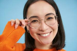 Portrait of smiling beautiful woman in glasses, looks happy at camera, standing over blue studio background photo