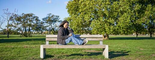 Young woman sitting alone on bench in park, using mobile phone, looking at screen, texting, sending messages in app chat photo
