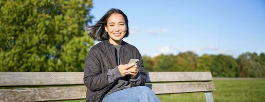 Technology and people. Young woman using smartphone, sitting outdoors on bench in park, looking at smartphone, sending message or networking photo