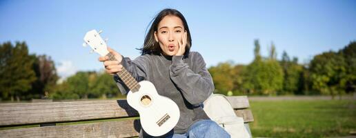 retrato de joven niña músico, sentado en parque con ukelele guitarra, mirando sorprendido a cámara, diciendo Guau foto