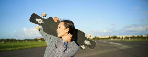 Happy and free asian girl holding cruiser board on shoulders and walking towards camera on empty road, skating on longboard and enjoying sunny weather photo