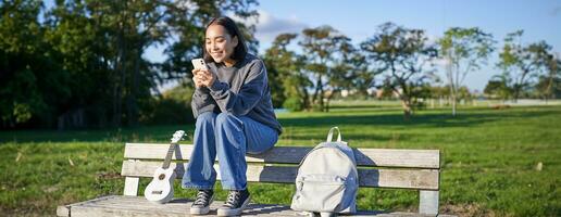 joven mujer sentado en parque en banco con ukelele, mirando a teléfono inteligente, leyendo mensaje en móvil teléfono y sonriente foto
