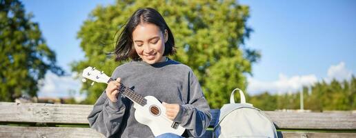 hermosa asiático niña obras de teatro ukelele al aire libre, se sienta en parque en banco con musical instrumento foto