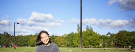 Vertical shot of cute teen girl sits in park on grass with backpack and her book, reading alone outdoors photo