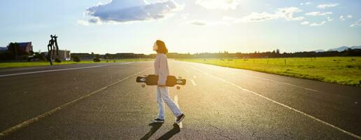 Young skater girl, teenager skating on cruiser, holding longboard and walking on concrete empty road photo