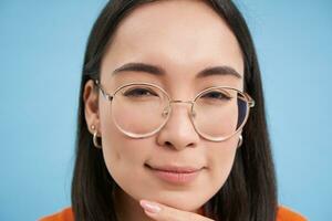 Close up portrait of asian woman looks intrigued, wears glasses, squints thoughtful, thinking, making assumption, standing over blue background photo