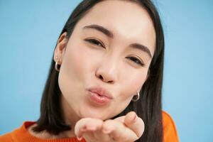 Close up of beautiful korean woman, blowing air kiss at camera, standing over blue background photo