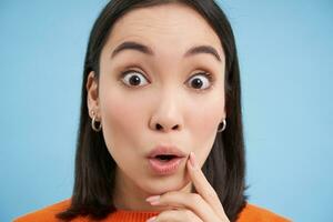 Close up portrait of girl looks with surprised, amazed eyes, says wow, impressed by smth, stands over blue background photo