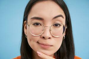 cerca arriba retrato de asiático mujer mira intrigado, usa anteojos, bizquear considerado, pensamiento, haciendo suposición, en pie terminado azul antecedentes foto