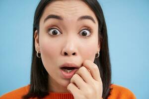 Close up portrait of amazed asian girl, looks with interest, listens to amazing story, impressed by something, stands over blue background photo