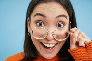 Close up portrait of amazed girl, looks closer at camera in glasses, standing against blue background photo