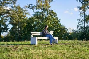 Side view of young woman sitting alone in park on bench, using her laptop to study or work remotely from outdoors photo