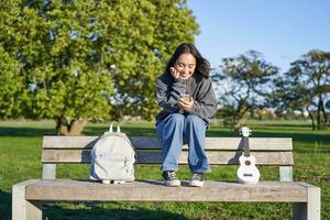 joven mujer con ukelele, sentado en banco en parque, utilizando móvil teléfono aplicación, participación teléfono inteligente y sonriente foto