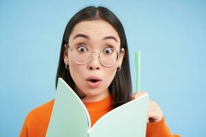 Amazed young asian woman in glasses, has an idea, writes down with pen in notebook, has revelation, enjoys listening course, student studying, blue background photo