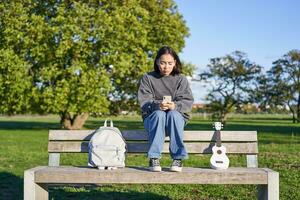 hermosa morena niña en banco en parque, sentado con ukelele y mochila, participación teléfono inteligente, utilizando móvil aplicación foto