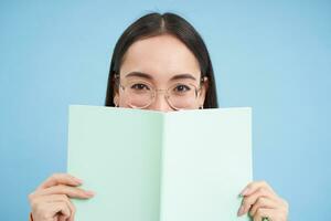 Portrait of cute japanese woman in glasses, peeking from notebook, covers face with her notes, smiles with eyes, blue background photo
