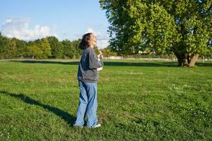 contento joven mujer sensación despreocupado, caminando en verde parque y jugando ukelele, canto a lo largo foto