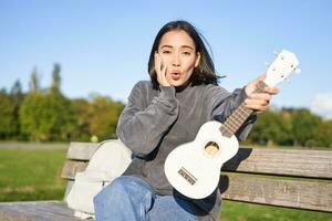 Portrait of young girl musician, sitting in park with ukulele guitar, looking surprised at camera, saying wow photo
