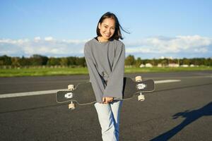 Portrait of beautiful asian girl skating on longboard, crusing with skateboard on empty road, enjoying freetime on fresh air photo