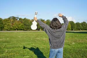 Upbeat young woman dancing with her musical instrument. Girl raises her ukulele up and pose in park on green field photo