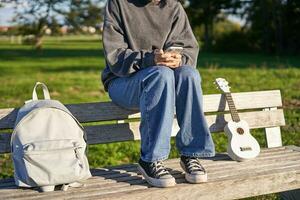 recortado Disparo de adolescente niña cuerpo, sentado en banco con ukelele, utilizando teléfono inteligente, manos participación móvil teléfono foto