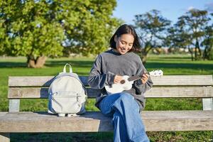 hermosa asiático niña obras de teatro ukelele al aire libre, se sienta en parque en banco con musical instrumento foto