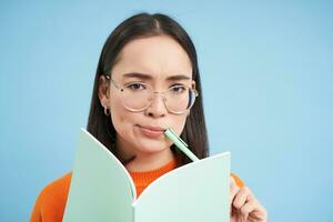 Portrait of asian girl, student in glasses, thinking, writing homework, making notes, holding pen, frowns and looks puzzled, blue background photo