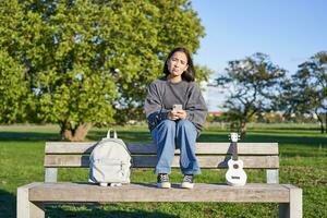 Girl with sad face, sitting on bench with smartphone and ukulele, looking upset and disappointed, being alone in park photo