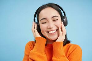 cerca arriba de hermosa asiático mujer en auriculares, sonrisas y escucha música, disfruta podcast, soportes terminado azul antecedentes foto