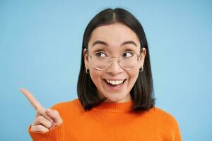 Portrait of japanese woman in glasses, points left, looks aside at banner, stands over blue studio background photo