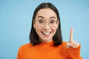 Portrait of smiling brunette asian woman in glasses, raises one finger, eureka sign, pitching an idea, has revelation, stands over blue background photo