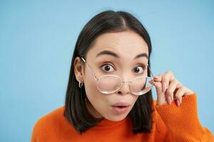retrato de asiático mujer en anteojos, mirando sorprendido, vistiendo gafas y curioso con interesar y curiosidad a cámara, azul antecedentes foto