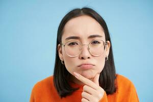 Close up of asian woman in glasses, thinking, staring at camera with thoughtful face expression, standing over blue background photo