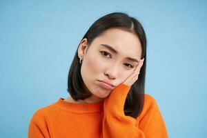 Close up of sad and gloomy japanese girl, sulking and looking upset, stands over blue background photo