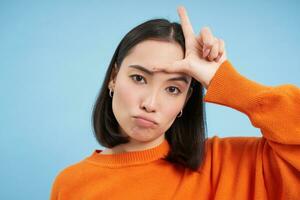 Close up of sassy korean girl, shows letter L, loser symbol on forehead, stands over blue studio background photo