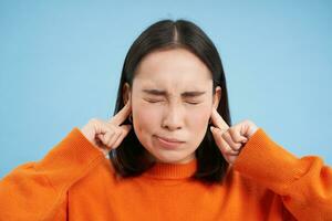 Close up of young woman shuts her ears with fingers and closes eyes, annoyed with loud noise, irritating sound, stands over blue background photo