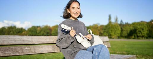 Happy cute girl sits alone on bench in park, plays ukulele guitar and enjoys sunny day outdoors photo