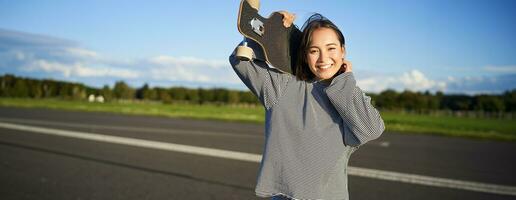 retrato de hermosa joven patinador chica, en pie con longboard y sonriente a cámara. asiático mujer con patineta en pie en la carretera foto