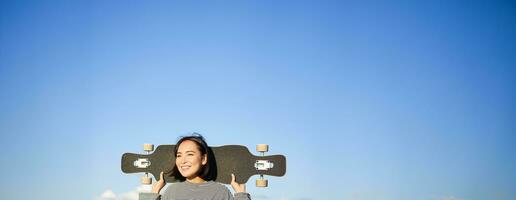 Vertical shot of carefree asian girl with longboard. Young woman skater holding cruiser on her shoulders and walking on road, skateboarding photo