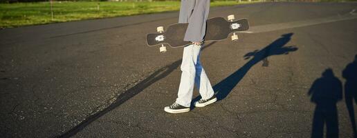 Cropped shot of teen girl body, holding cruiser longboard in hand, walking in sneakers on road in jeans and sweatshirt. Young woman skater with skateboard photo