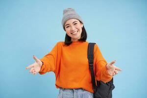 Friendly Korean woman in hat with backpack, reaches hands for hug, receives smth, leans to cuddle you, warm welcome, stands over blue background photo