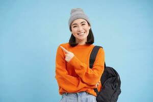 Beautiful smiling korean woman in hat, pointing finger left, showing information banner, demonstrates offer, stands over blue background photo