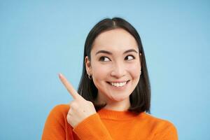 Close up shot of smiling asian girl, points finger at upper left corner, shows banner, demonstrates advertisement, blue background photo