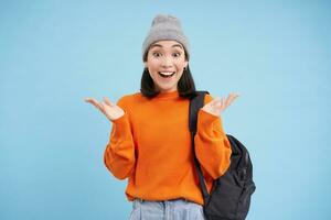 Happy asian woman in warm hat, claps hands from surprise, laughs and looks amazed, excited by smth, stands over blue background photo